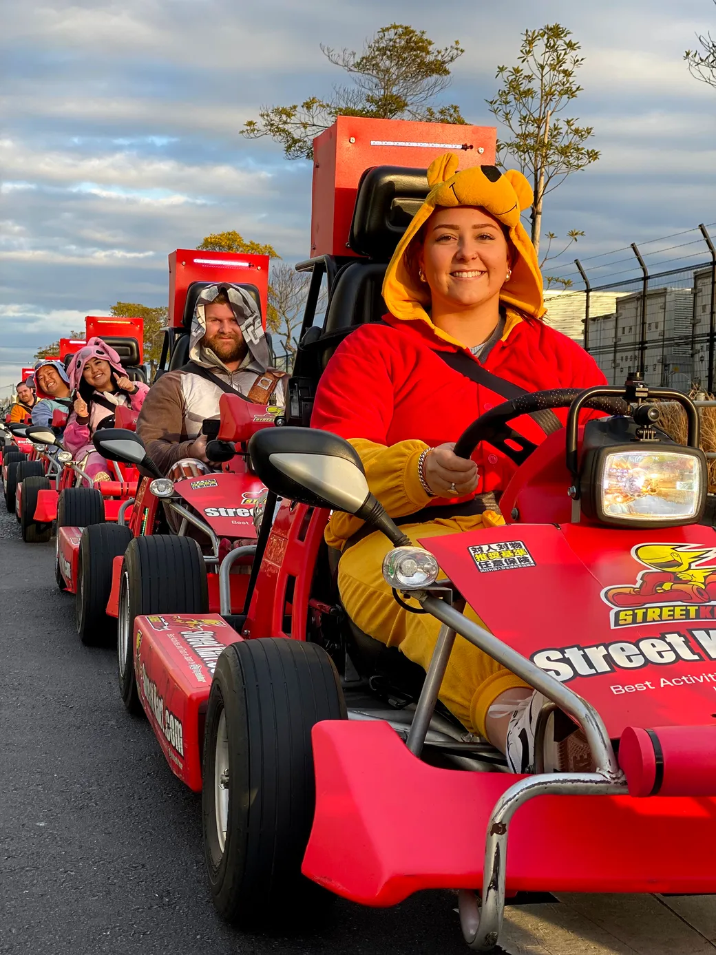 Smiling woman in a yellow bear costume drives a red go-kart at the front of a line of riders in matching red karts.
