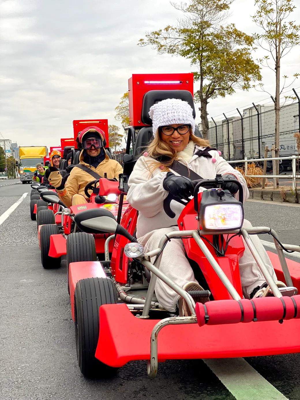 Woman in a white coat and knit hat drives a bright red go-kart on a city street, with other riders in line behind her.