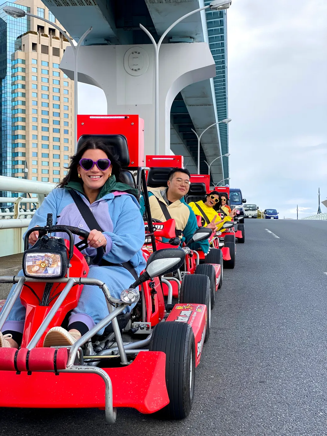 Group of people riding bright red go-karts along an urban bridge, front rider in a blue jacket and purple sunglasses smiling at the camera.